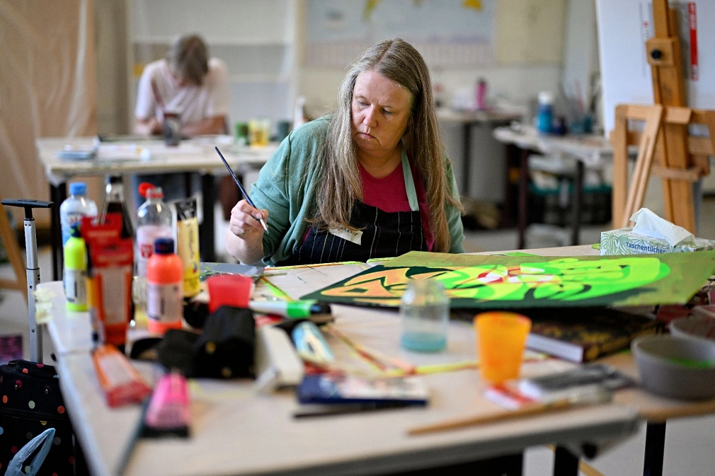 Sommerakademie: Kurs Konstantin Voit, Schablonenmalerei Kursteilnehmerin arbeitet an einem Bild, auf dem Tisch vor ihr stehen Malutensilien und Farben. © Georg Kronenberg