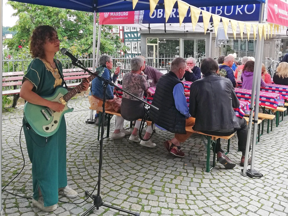 Im Vordergrund steht eine Frau an einem Mikrofon. Sie hält eine Gitarre. Im Hintergrund sind Sitzbänke an Tischen aufgestellt. Auf den Bänken sitzen Menschen.