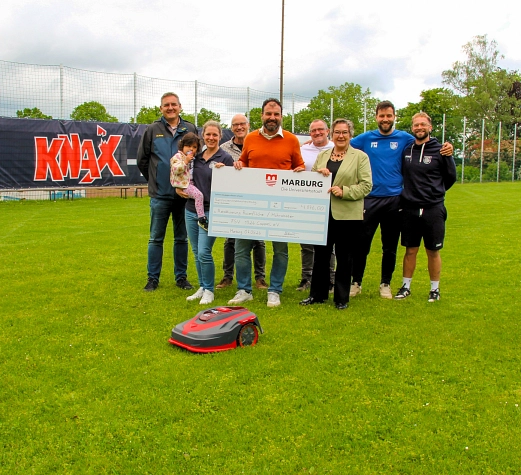 Menschen stehen in einer Gruppe auf einem Fußballplatz. Sie halten einen großen Scheck in der Hand.