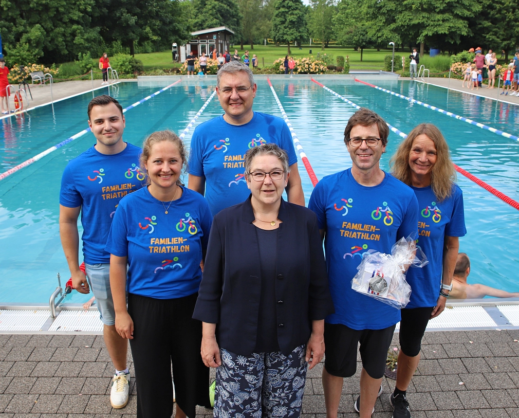 Auf dem Bild sieht man 6 Personen, die vor einem Schwimmbecken stehen. Eine der vorderen Personen wurde geehrt und trägt ein kleines Geschenk in ihren Händen.