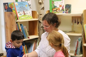 Eine Mitarbeiterin der Stadtbücherei hält ein Buch in den Händen. Sie kniet auf dem Boden vor zwei Bücherregalen. Vor ihr sitzt ein Junge auf einem Hocker, ein Mädchen kniet neben ihr. Beide Kinder sehen auf die Titelseite des Buches, auf die die Mitarbeiterin deutet.