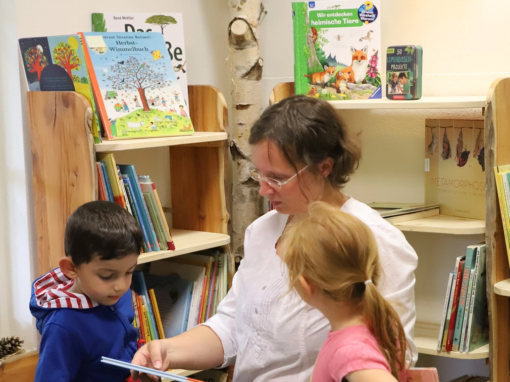 Eine Mitarbeiterin der Stadtbücherei hält ein Buch in den Händen. Sie kniet auf dem Boden vor zwei Bücherregalen. Vor ihr sitzt ein Junge auf einem Hocker, ein Mädchen kniet neben ihr. Beide Kinder sehen auf die Titelseite des Buches, auf die die Mitarbeiterin deutet.&nbsp;&copy;&nbsp;Stefanie Profus, i.A.d. Stadt Marburg