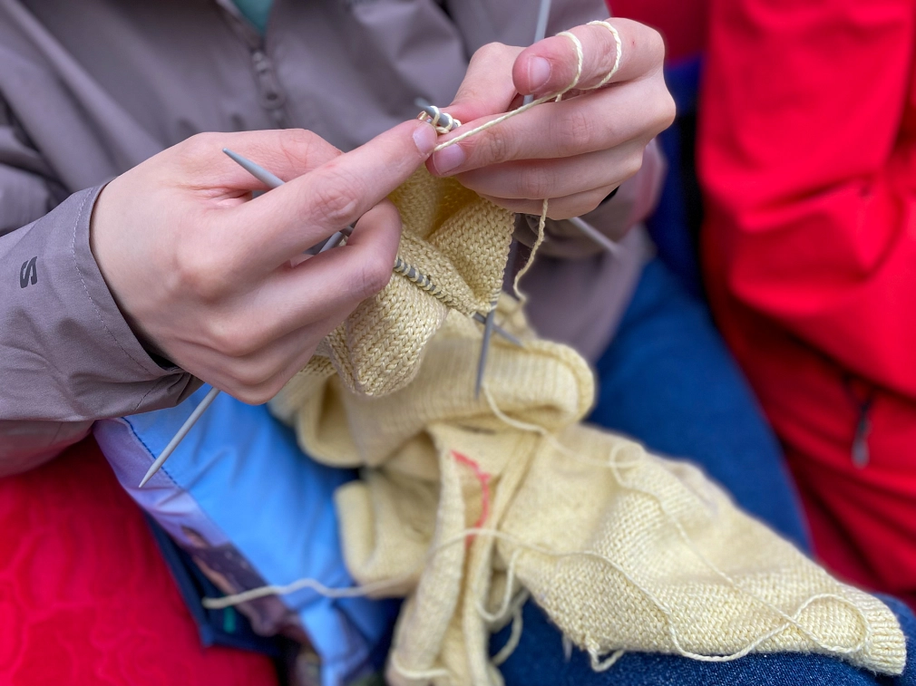 Zwei Hände halten Stricknadeln in der Hand und stricken mit gelber Wolle ein Oberteil.