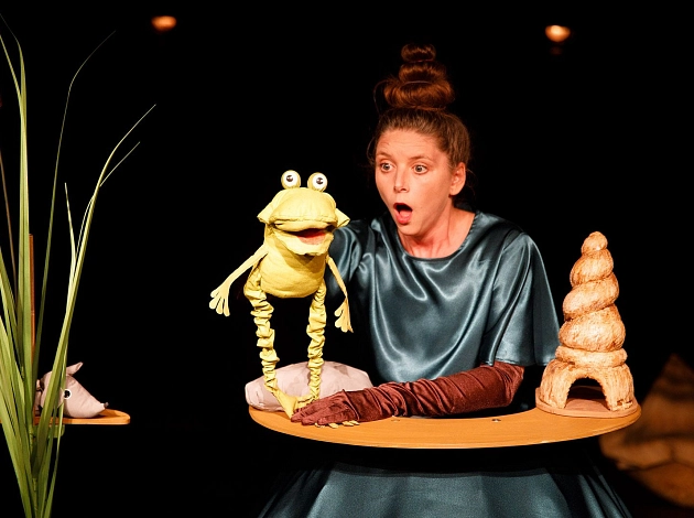 Eine Frau im blauen Kleid sitzt an einem Tisch auf einer Bühne, sie guckt erstaunt. Sie hat einen Frosch als Handpuppe.