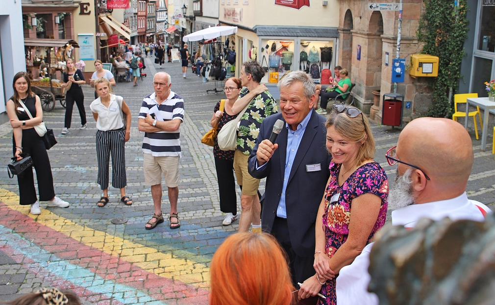 Das Bild zeigt einen Mann mit Mikrofon in der Hand, der zu einer Menschenmenge spricht, welche in einer Kopfsteinpflaster-Straße stehen. Auf der Straße ist ein Regenbogen gemalt.