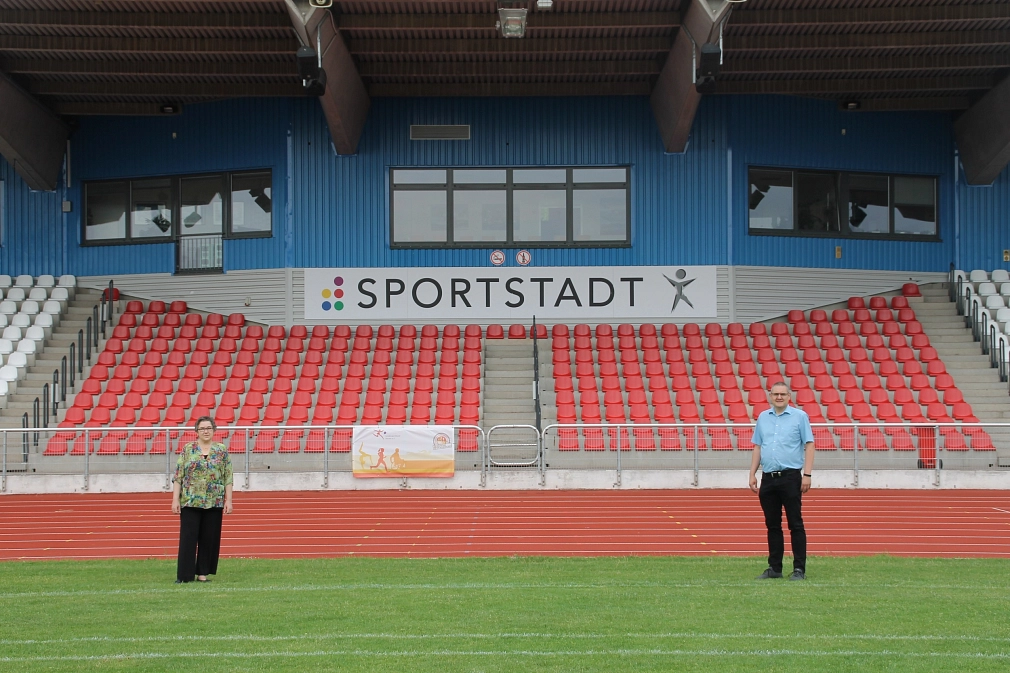 Rote und weiße Sitzschalen im Georg-Gaßmann-Stadion, im Hintergrund der Tribüne hängt ein Schild mit der Aufschrift "Sportstadt". Im Vordergrund stehen Stadträtin Kirsten Dinnebier und Fachdienstleiter Björn Backes.