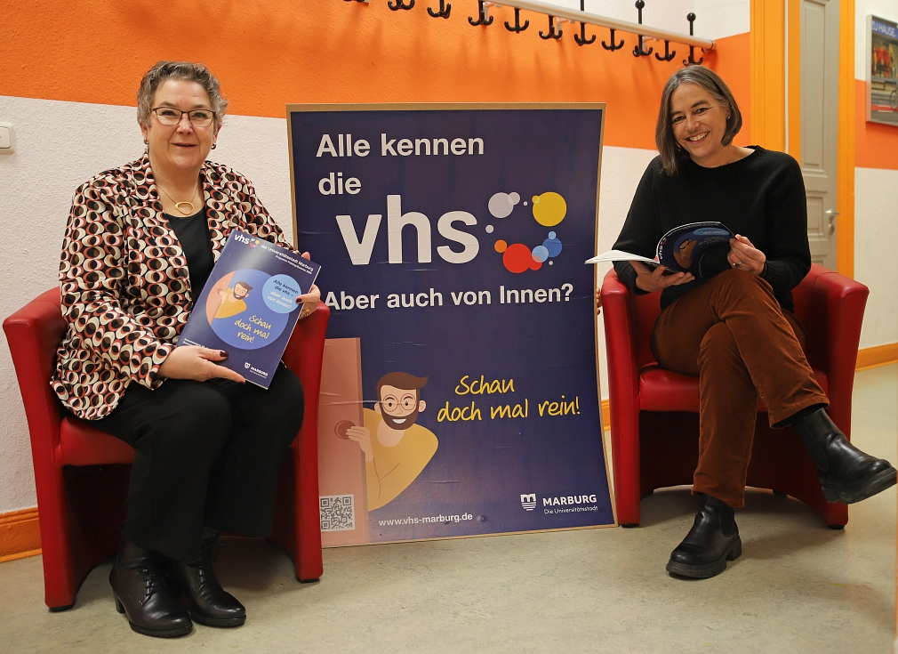Zwei Frauen sitzen auf zwei roten Sesseln. Zwischen Ihnen ist ein Plakat mit dem Logo der vhs abgebildet. Beide halten ein Heft in der Hand.