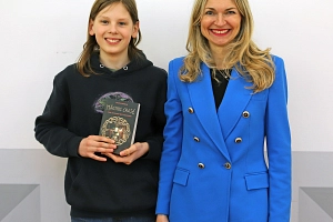 Vorlesewettbewerb 2026 Zwei Personen stehen vor einer Wand. Beide lächeln in die Kamera. Der Junge hält ein Buch in der Hand.