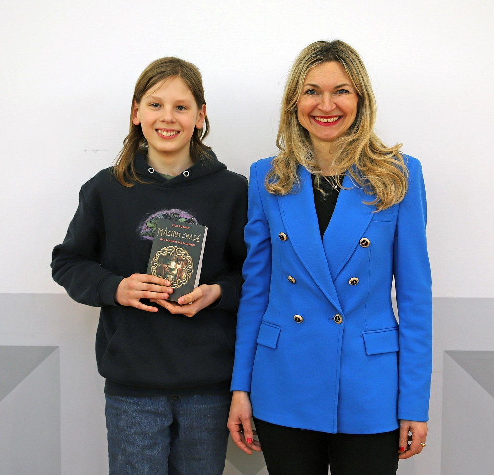 Zwei Personen stehen vor einer Wand. Beide lächeln in die Kamera. Der Junge hält ein Buch in der Hand.