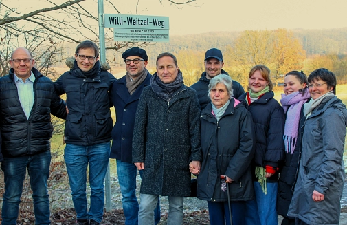 Eine Gruppe Erwachsener stehen unterhalb des Straßenschildes am Rande eines Weges.
