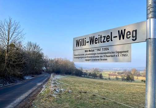 Straßenschild mit der Aufschrift "Willi-Weitzel-Weg" und darunter ein Schild mit folgender Info: Willi Weitzel, geboren 1941, gestorben 2024, Ehrenortsvorsteher, jahrzehntelanges kommunalpolitisches Engagement, Gründungsmitglied und Ehrenvorsitzender des SV Bauerbach e.V. (1965), engagiert in weiteren Vereinen.