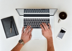Aus der Vogelperspektive fotografiert zwei Hände, die einen weißen Laptop bedienen, daneben ein Smartphone, eine Tasse Kaffee und ein Notizbuch © stokpic / Pixabay